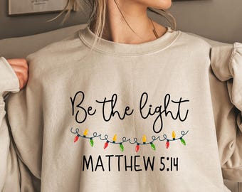 May include: Beige sweatshirt with the text "Be the light" in black script, with a string of colorful Christmas lights underneath. The Bible verse Matthew 5:14 is printed below the lights.