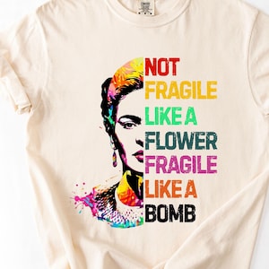 以下が含まれることがあります： 女性の顔のカラフルなグラフィックと、さまざまな色の「NOT FRAGILE LIKE A FLOWER FRAGILE LIKE A BOMB」というテキストが特徴のクリーム色のTシャツ。 このデザインには、鮮やかな水彩効果が含まれています。