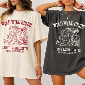 Op de afbeelding: Twee oversized T-shirts, één crème en één antracietgrijs, met een grafisch ontwerp. Het crèmekleurige shirt leest "Wild Wild Bride" en het grijze "Wild Wild Crew". Beide shirts hebben cowboylaarzen, een cocktail en "Lena's Bachelorette, Fredericksburg, TX."