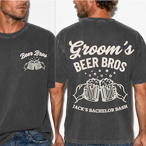May include: A charcoal gray short-sleeve t-shirt featuring cream-colored graphics. The front displays "Beer Bros" with two clinking beer mugs. The back features "Groom's BEER BROS" and "JACK'S BACHELOR BASH" with a larger illustration of clinking beer mugs.