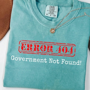 Puede incluir: Una camiseta Comfort Colors turquesa con el texto "ERROR 404 Government Not Found!" en rojo y blanco. Un collar dorado con un colgante está sobre la camiseta. La camiseta es talla S.