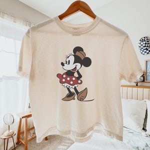 May include: Cream-colored t-shirt featuring a classic Minnie Mouse graphic. The cartoon character is depicted in a red polka-dot skirt, brown shoes, and a matching hat. The shirt is hanging on a wooden hanger.