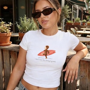 May include: A white short-sleeved t-shirt featuring a graphic of a brown monkey carrying a red surfboard. Below the graphic, the text reads, "I don't want this feeling to go away." The shirt has a crew neckline and a fitted silhouette.