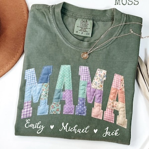 以下が含まれることがあります： モスグリーンのTシャツに、パッチワーク文字で「MAMA」の文字がデザインされています。文字は様々な模様と色で表現されています。その下に「Emily」、「Michael」、「Jack」の名前が書かれています。金のペンダントネックレスも写っています。