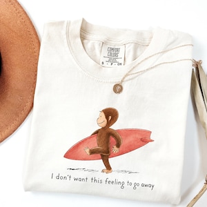 May include: A cream-colored t-shirt featuring a graphic of a brown monkey carrying a red surfboard. Below the graphic, the text reads, "I don't want this feeling to go away." The shirt has a Comfort Colors brand label at the inner neckline.