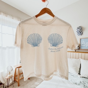 May include: Cream-colored t-shirt featuring two light blue seashell designs. The text "Sophie's Bachelorette Miami, 2026" is printed below the seashells. The shirt is hanging on a wooden hanger.