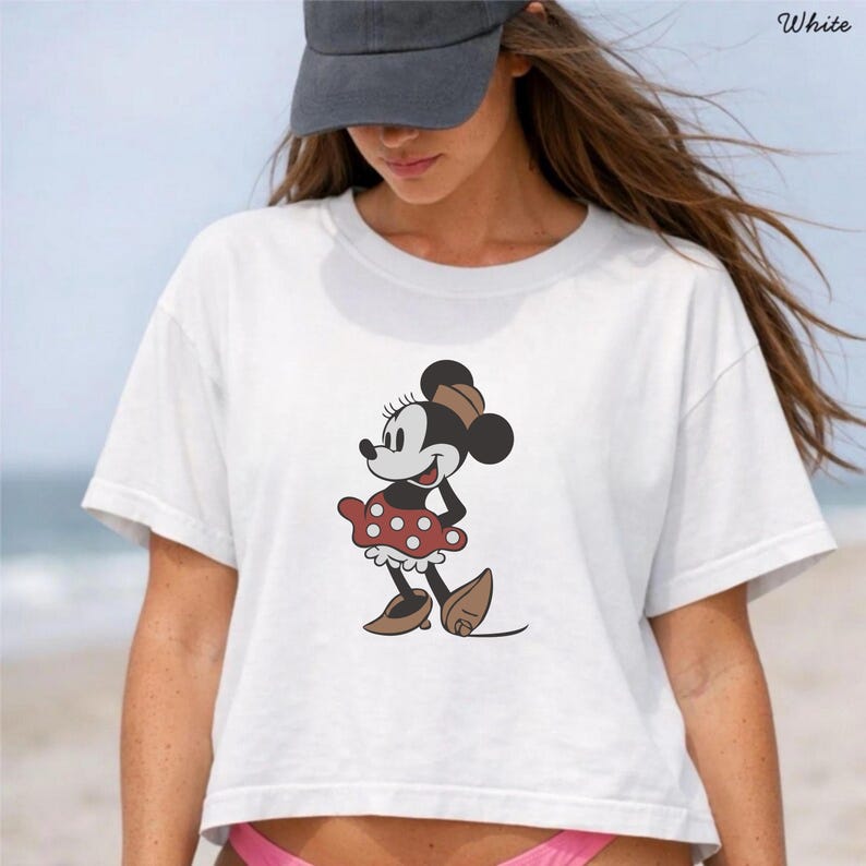 May include: White crop top featuring a Minnie Mouse graphic. The cartoon character is depicted in a classic pose, wearing a red polka-dot skirt, brown shoes, and a matching bow. The word "White" is printed in the upper right corner.
