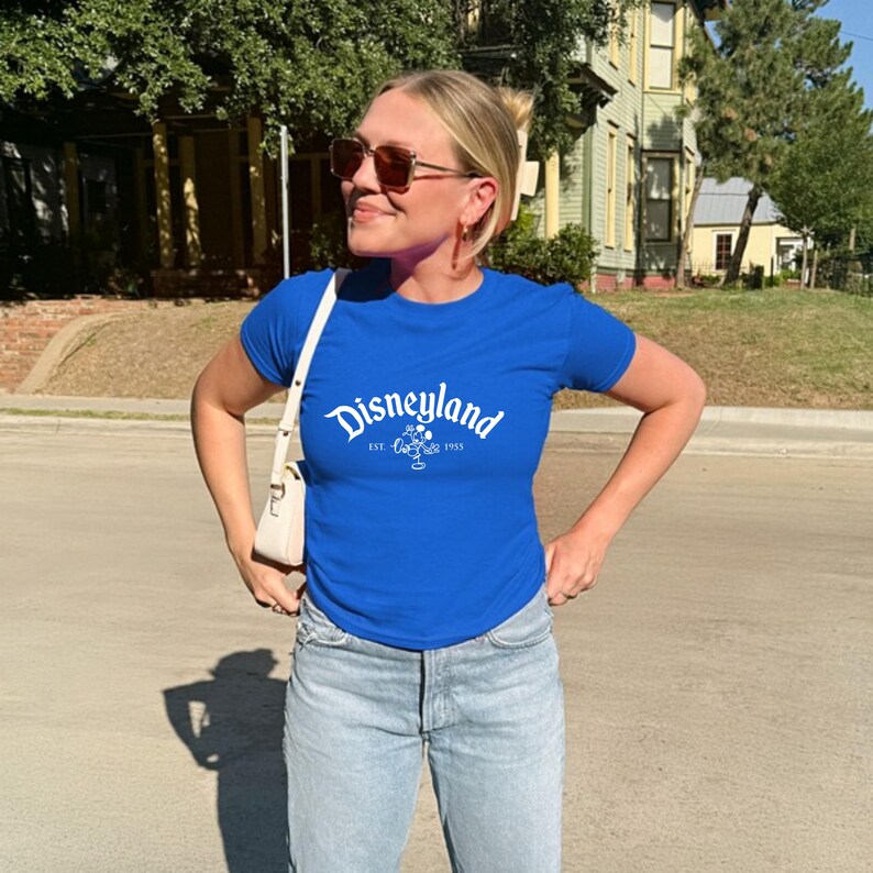 May include: A royal blue t-shirt with the word "Disneyland" in white script across the chest. The shirt also features a small graphic and the text "EST. 1955". The shirt is paired with light wash denim jeans.