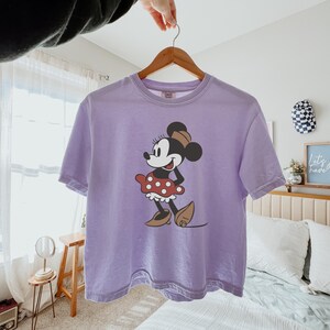 May include: A light purple t-shirt featuring a graphic of Minnie Mouse. Minnie is depicted in her classic outfit: a red polka-dot skirt, brown shoes, and a matching bow. The shirt is hanging on a wooden hanger.
