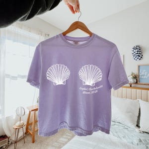 May include: A light purple t-shirt with a white seashell design and the text "Sophie's Bachelorette Miami, 2026". The shirt is hanging on a wooden hanger.