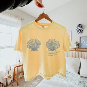 May include: Yellow t-shirt with two blue seashell graphics and the text "Sophie's Bachelorette Miami, 2026". The shirt is hanging on a wooden hanger.