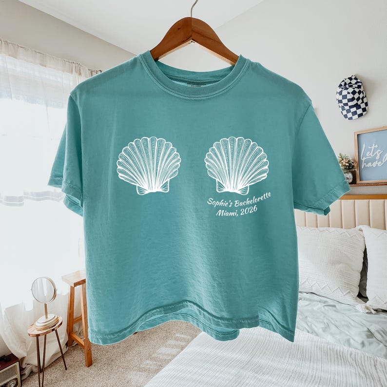 May include: Teal-colored t-shirt with two white seashell graphics. The text "Sophie's Bachelorette Miami, 2026" is printed below the shells. The shirt is hanging on a wooden hanger.