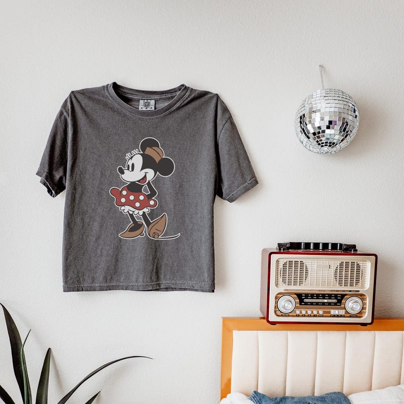 May include: A charcoal gray t-shirt featuring a graphic of Minnie Mouse in a red polka dot dress and brown shoes. The classic cartoon character is smiling. The shirt is short-sleeved and has a crew neck.