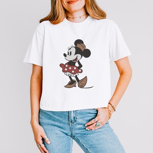 May include: White t-shirt featuring a cartoon illustration of Minnie Mouse. The character is depicted in a classic style, wearing a red polka-dot dress, brown shoes, and a matching bow. The shirt is a casual, everyday wear item.