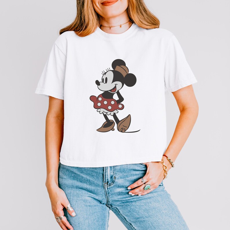 May include: White t-shirt featuring a cartoon illustration of Minnie Mouse. The character is depicted in a classic style, wearing a red polka-dot dress, brown shoes, and a matching bow. The shirt is a casual, everyday wear item.