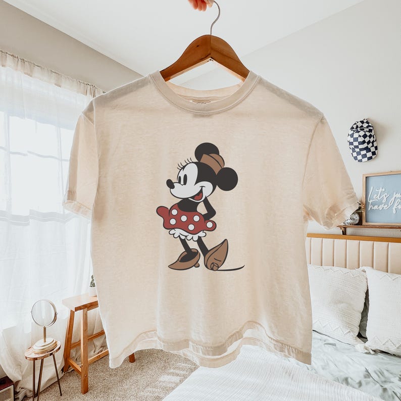 May include: Cream-colored t-shirt featuring a classic Minnie Mouse graphic. The cartoon character is depicted in a red polka-dot skirt, brown shoes, and a matching hat. The shirt is hanging on a wooden hanger.