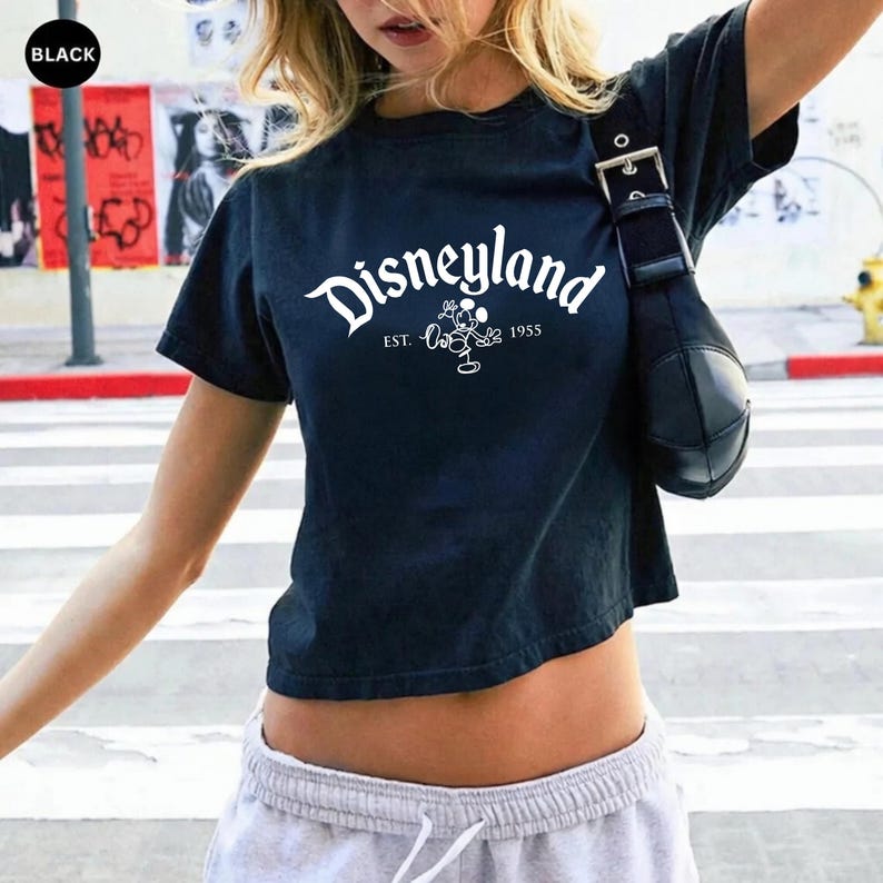 May include: Black cropped t-shirt with the word "Disneyland" in white script. Below the word is a graphic of Mickey Mouse and the text "EST. 1955". The shirt is short-sleeved.