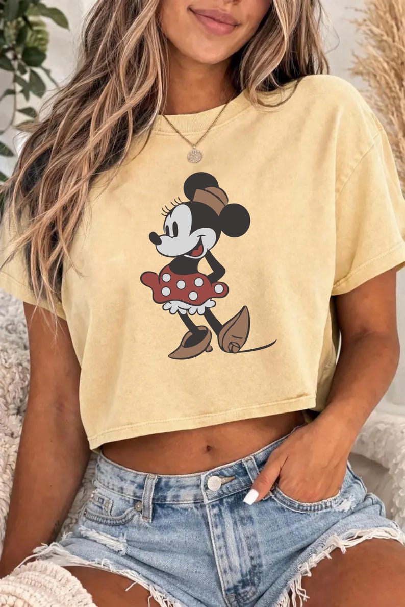 May include: Yellow crop top featuring a Minnie Mouse graphic. The cartoon character is depicted in a red polka-dot skirt, brown shoes, and a matching hat. The shirt is a light yellow color.