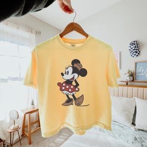May include: Yellow t-shirt featuring a graphic of Minnie Mouse. The cartoon character is depicted in a classic pose, wearing a red polka-dot skirt, brown shoes, and a matching bow. The shirt is hanging on a wooden hanger.