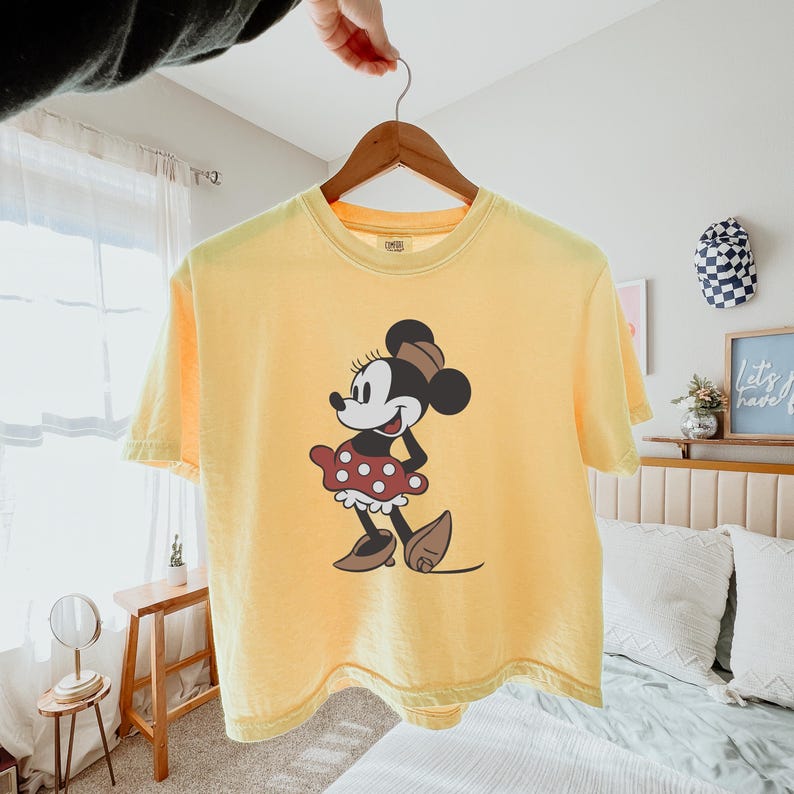 May include: Yellow t-shirt featuring a graphic of Minnie Mouse. The cartoon character is depicted in a classic pose, wearing a red polka-dot skirt, brown shoes, and a matching bow. The shirt is hanging on a wooden hanger.