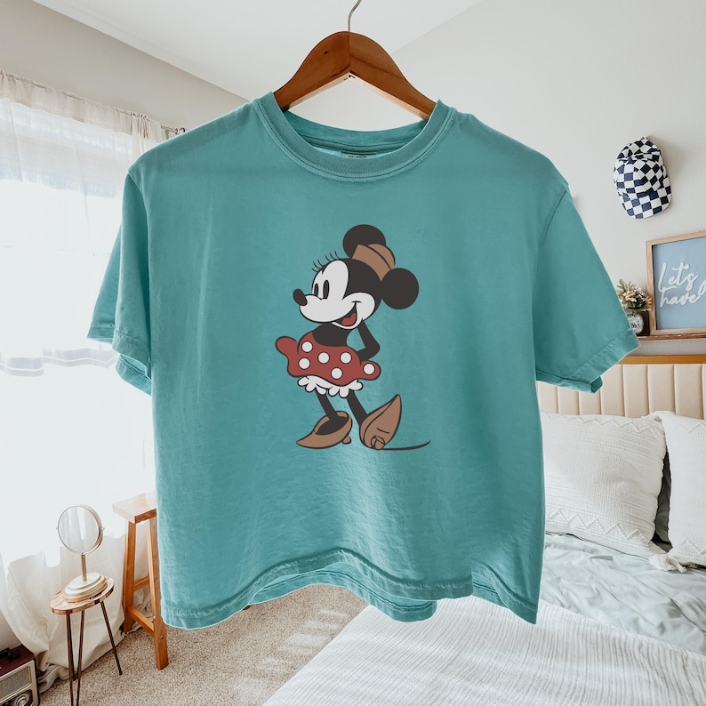 May include: Teal-colored t-shirt featuring a graphic of Minnie Mouse. Minnie is depicted in a classic pose, wearing a red polka-dot skirt, brown shoes, and a matching bow. The shirt is hanging on a wooden hanger.