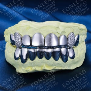 May include: Silver-colored grillz with diamond accents, displayed on a dental mold. The grillz feature a polished finish and are designed to cover the upper teeth. The image includes text that reads "EONLEAD FINE JEWELRY TOP CUSTOM REAL FACTORY PRICES."