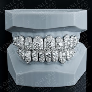 May include: A pair of diamond-encrusted grillz, presented on a grey dental model. The grillz are covered in numerous round and square-cut diamonds, creating a sparkling effect. The image includes the text "EONLEAD" and "FINE JEWELRY".