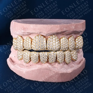 May include: Gold-colored, diamond-studded grillz set. The top and bottom teeth are fully covered in small, sparkling diamonds. The grillz are displayed on a pink dental mold, with text in the background.