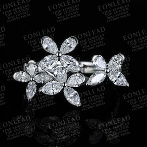 Könnte beinhalten: Ein silberfarbener Ring mit einer Ansammlung von klaren, birnenförmigen und marquise-geschliffenen Steinen, die in Blumenmustern angeordnet sind. Das Design umfasst zwei blumenartige Gruppierungen auf einem Metallring vor dunklem Hintergrund.