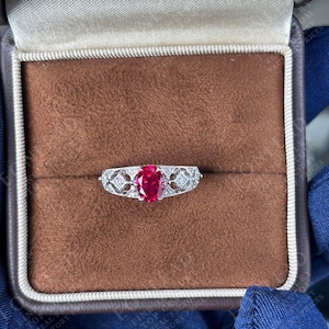 Op de afbeelding: Een zilveren ring met een levendige rode ovaalvormige edelsteen in het midden. De band heeft ingewikkelde filigraan- en diamantaccenten. De ring wordt gepresenteerd in een bruine sieradendoos met een fluwelen binnenkant.