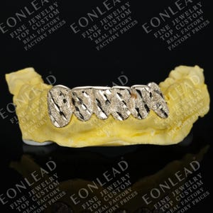 May include: Gold-colored custom grillz with a textured, diagonal pattern. The grillz are displayed on a yellow dental mold. The image includes the text "EONLEAD FINE JEWELRY TOP CUSTOM REAL FACTORY PRICES".