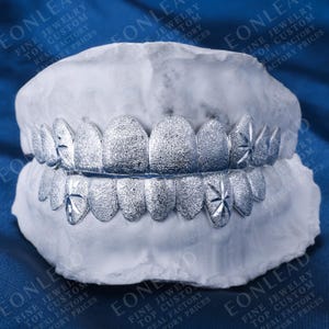 May include: Silver-colored grillz with a textured surface and starburst designs on the canines and incisors. The grillz are displayed on a white dental mold. The image has the text "EONLEAD" and "FINE JEWELRY TOP CUSTOM REAL FACTORY FACTORY PRICES".