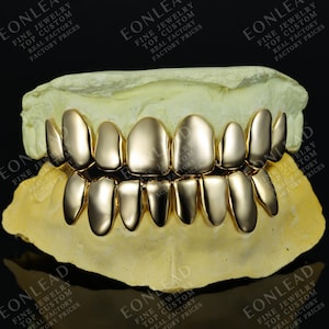 May include: Gold-colored dental grillz, displayed on a dental mold. The grillz are highly polished and reflect light. The image includes the text "EONLEAD FINE JEWELRY TOP CUSTOM REAL FACTORY FACTORY PRICES".