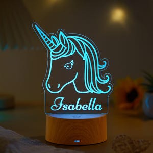 May include: A blue LED unicorn night light with the name "Isabella" etched on the front. The unicorn design is illuminated with a soft blue glow and sits on a wooden base. The light is designed for a child's room.