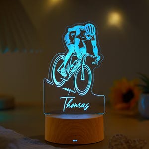 Può includere: Lampada in acrilico illuminata di blu raffigurante un ciclista in bicicletta, con il nome "Thomas" scritto sotto. La lampada è posta su una base di legno. Adatta agli appassionati di ciclismo.