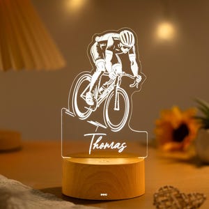 Peut inclure: Une lampe de nuit décorative avec un cycliste blanc sur un vélo, gravé sur un panneau acrylique transparent. Le nom "Thomas" est inscrit en dessous. La base est en bois rond et émet une lumière chaude.