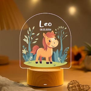 Puede incluir: Una luz de noche decorativa con un diseño de caballo de dibujos animados. Tiene una base de madera y un panel acrílico transparente con el nombre "Leo" y la fecha "08.05.2024" impresos. El caballo es naranja y marrón, sobre un fondo de árboles y follaje.