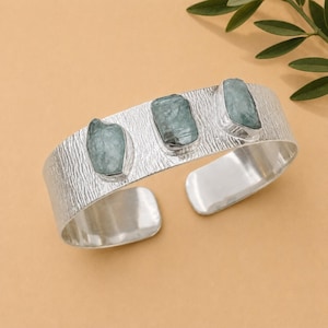 May include: A silver cuff bracelet featuring a textured, bark-like surface and three rectangular, raw, pale blue-green aquamarine stones set horizontally along the center of the band. The metal has a bright, polished finish on the interior.