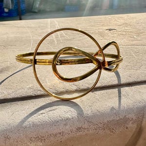 May include: A gold-colored metal bracelet with an abstract design. The bracelet features a circular element and an intertwined knot-like shape. The metal has a slightly aged appearance, and the bracelet is resting on a light-colored surface.