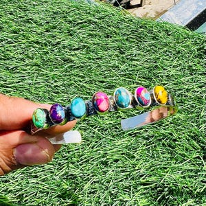 May include: A silver cuff bracelet featuring seven oval-shaped gemstones in various colors, including green, purple, turquoise, pink, and yellow. The gemstones are set in individual silver bezels, creating a vibrant and eye-catching design. The bracelet is displayed against a backdrop of green grass.