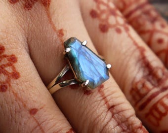 Könnte beinhalten: Ein silberner Ring mit einem rechteckigen, irisierenden blauen Labradorit-Edelstein. Das Band des Rings teilt sich an der Basis des Steins in zwei Teile, und kleine silberne Zinken sichern den Edelstein. Der Ring wird auf einer Hand mit Henna-Designs präsentiert.