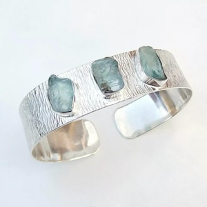 May include: A silver cuff bracelet featuring three light blue aquamarine stones. The bracelet has a textured surface and an open design. The aquamarine stones are set in silver bezels.