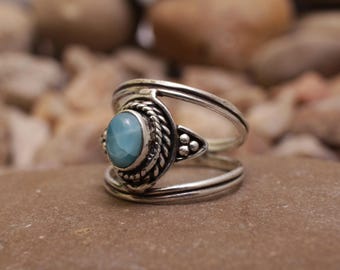 Può includere: Un anello d'argento con una pietra preziosa ovale azzurra. L'anello presenta un motivo intrecciato attorno alla gemma e un gambo diviso. L'anello è appoggiato su una superficie marrone.