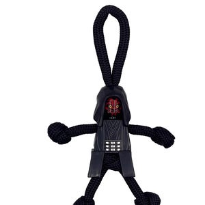 Puede incluir: Una figura negra de Darth Maul hecha de cordón trenzado, con una cara roja y negra, capucha negra y una placa de pecho detallada. La figura tiene brazos y piernas del mismo cordón, con un lazo en la parte superior para colgar.