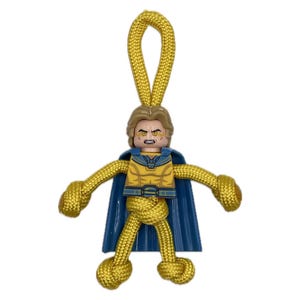 May include: A handmade keychain featuring a Lego figure with a blue cape and yellow and blue torso. The figure's arms and legs are crafted from knotted yellow cord, with a loop at the top for attachment. The figure has a stern expression.