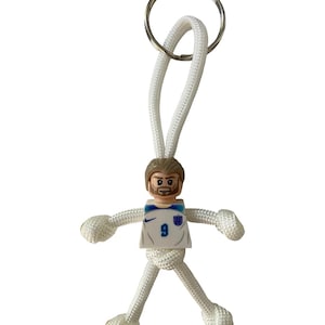 May include: A white keychain featuring a small figure made of a Lego head and a white jersey with the number 9. The figure has white rope arms and legs, and a silver key ring at the top.
