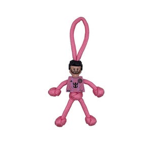 Op de afbeelding: Een roze handgemaakte sleutelhanger met een Lego-achtig figuur met zwart haar en een baard. Het figuur draagt een roze shirt met een teamlogo. De armen en benen zijn gemaakt van roze touw en de sleutelhanger heeft een lus.