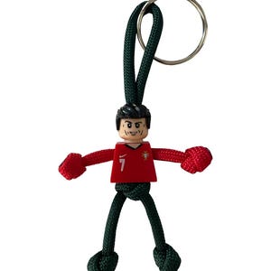 May include: A keychain featuring a small figure of a soccer player. The figure wears a red jersey with the number 7 and a green and red lanyard. The keychain has a silver ring.
