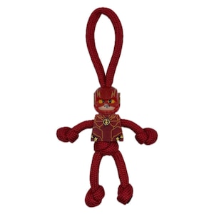 Może przedstawiać: Czerwony brelok do kluczy z motywem superbohatera Flasha. Brelok przedstawia małą figurkę Flasha w czerwono-złotym kostiumie i z emblematem błyskawicy. Figurka jest przymocowana do pętli z czerwonej liny.