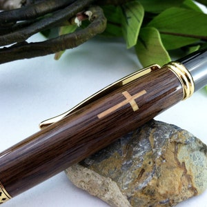 Könnte beinhalten: Ein Holzstift mit goldener Verzierung und einer Kreuz-Einlage. Der Stift hat eine schwarze Metallspitze und eine goldene Banderole um den Schaft.